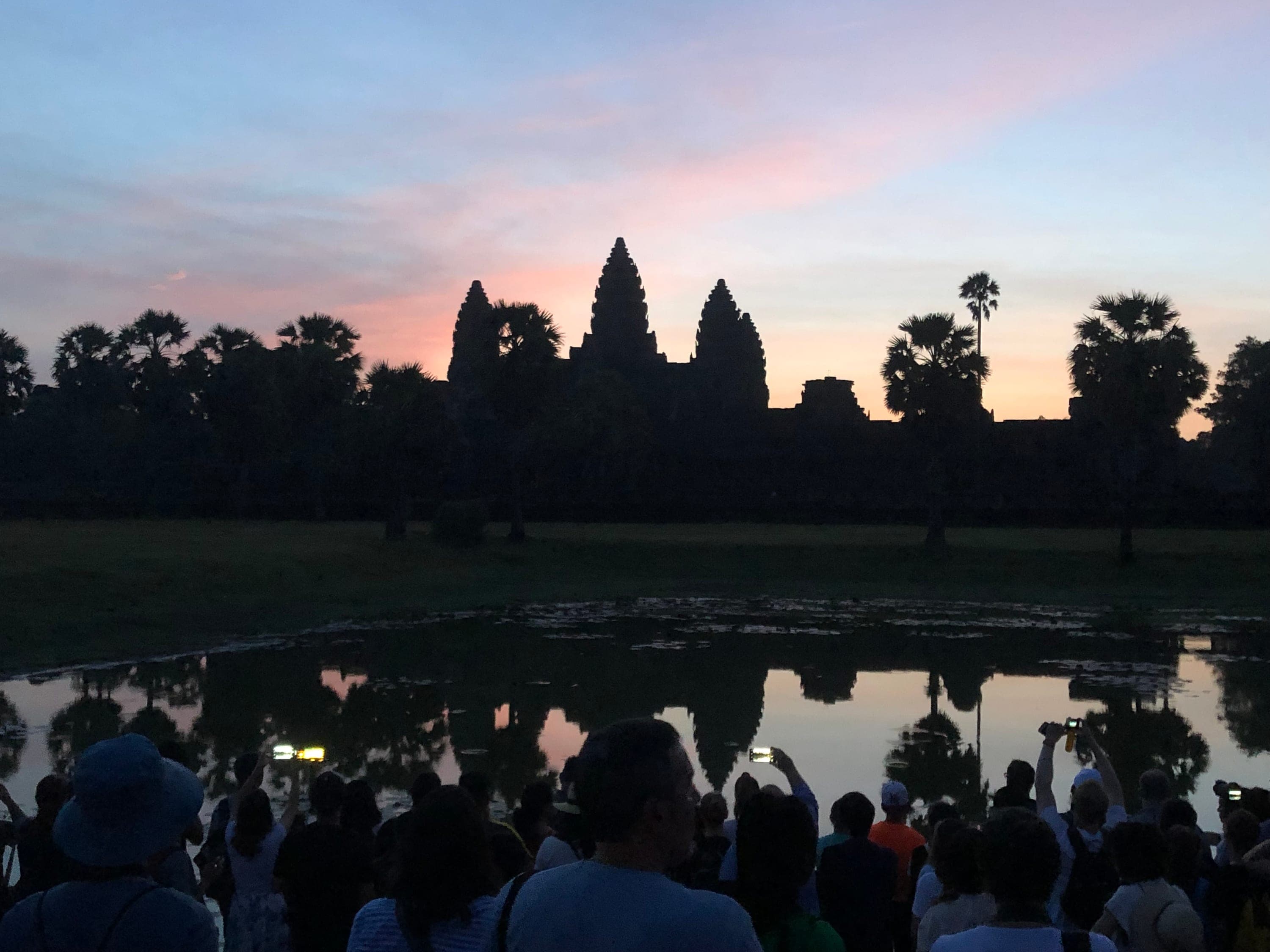 Angkor Wat at sunrise, temple silhouettes reflected in the lotus pond, viewers gathered along the bank.